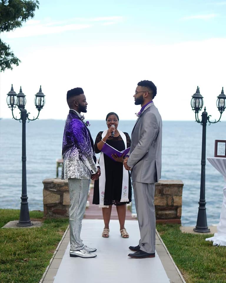 Two men in formal attire holding hands at an outdoor wedding ceremony by the water, with an officiant speaking to them.