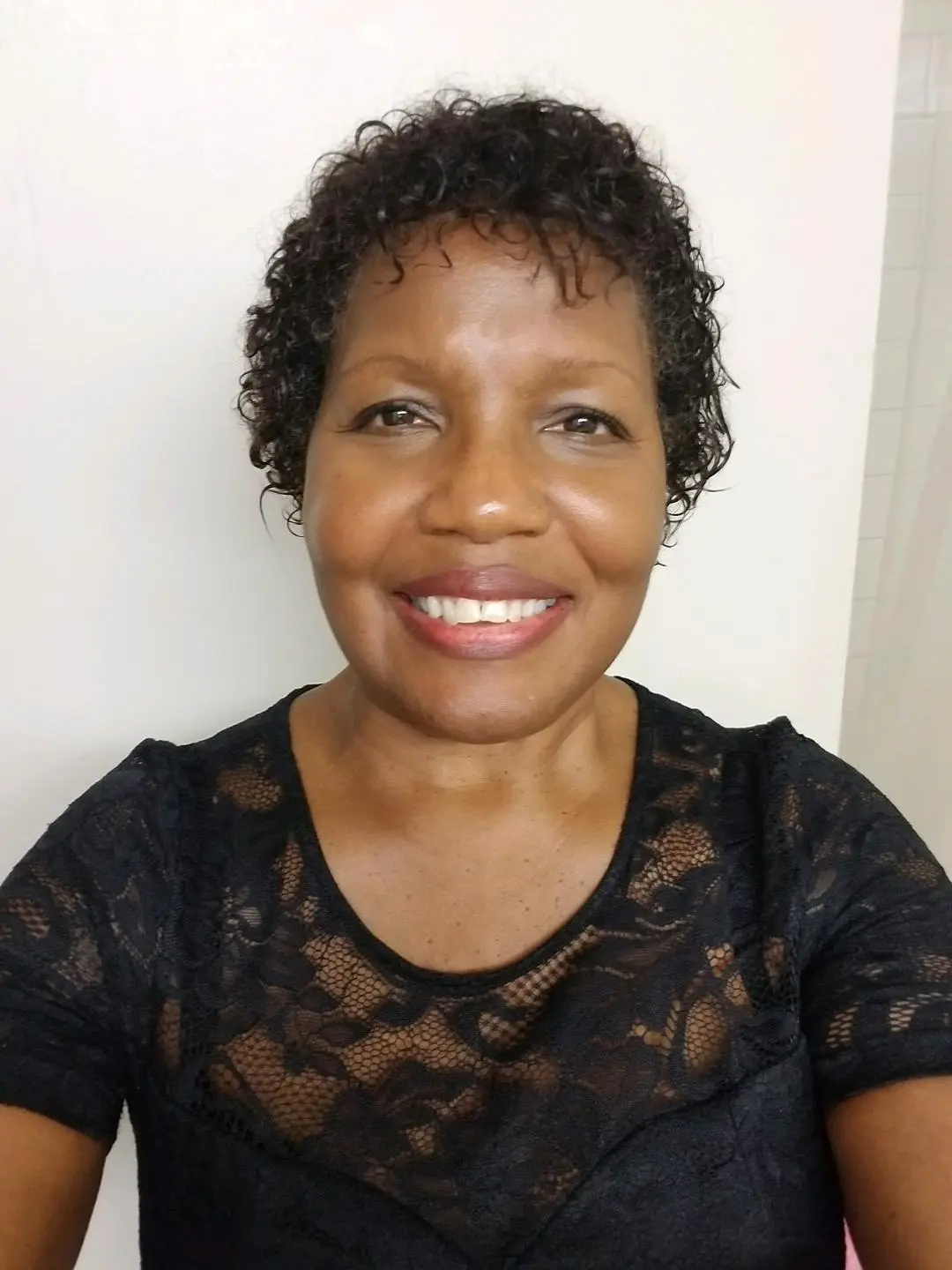 A smiling woman with short curly hair wearing a lace top, posing for a close-up photo in a white room.